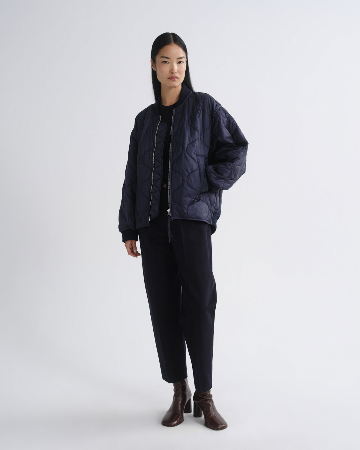 A model stands against a light gray studio background wearing a dark navy quilted bomber jacket over a black top, black trousers, and dark brown heeled ankle boots.