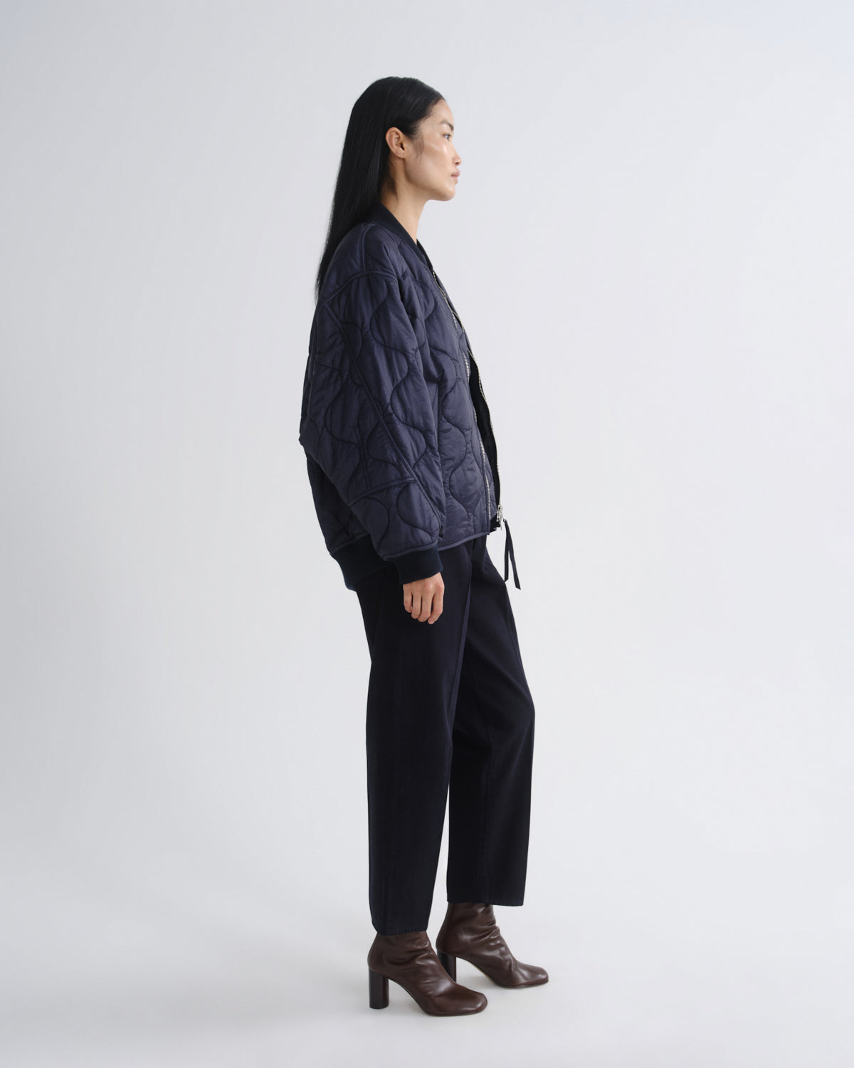 A woman stands in profile wearing a dark navy quilted jacket, black wide-leg trousers, and brown heeled ankle boots against a plain light gray studio background.