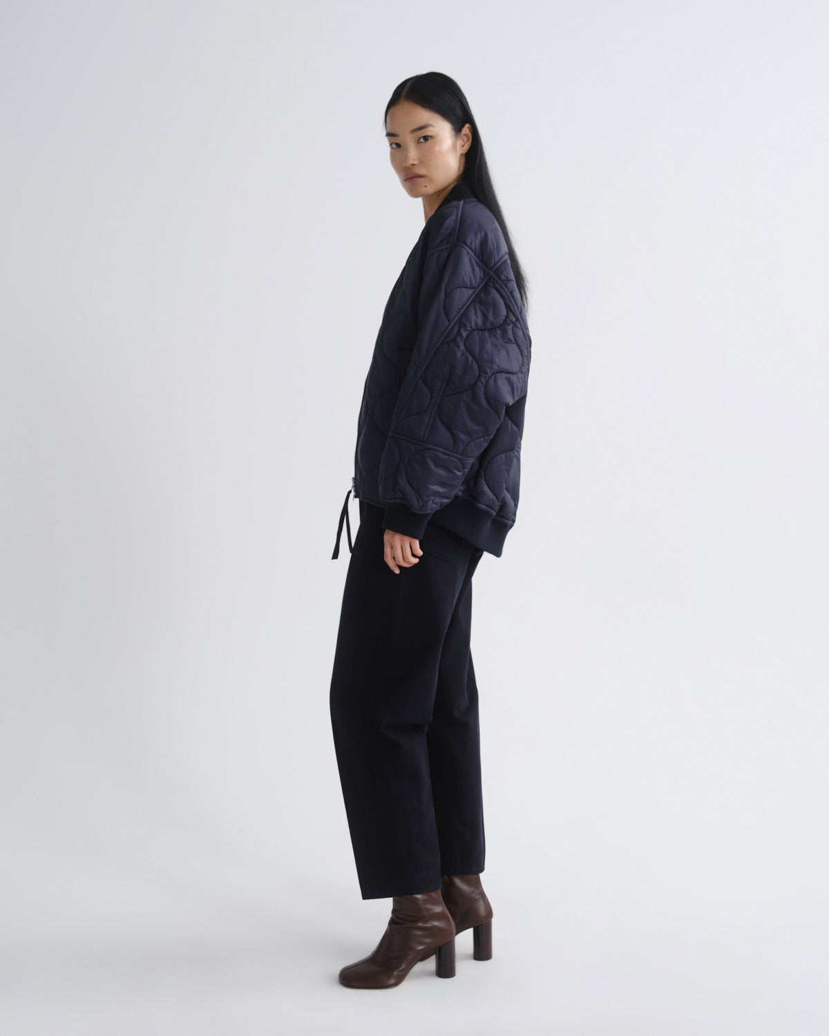 A model is shown in profile wearing a dark navy quilted jacket, black wide-leg trousers, and brown heeled ankle boots against a plain light gray studio background.