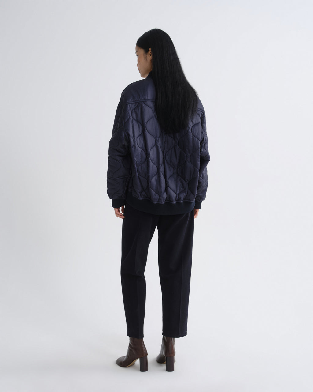 A model viewed from the back wearing a dark navy quilted puffer jacket, black trousers, and brown heeled ankle boots against a plain light gray studio background.