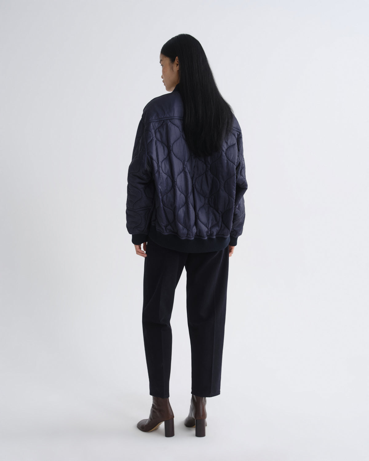 A model viewed from the back wearing a dark navy quilted puffer jacket, black trousers, and brown heeled ankle boots against a plain light gray studio background.