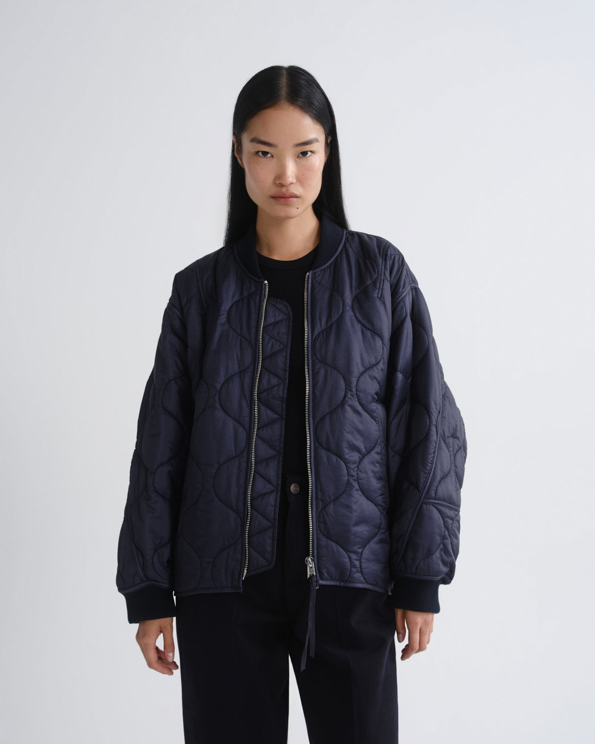 A model wearing a dark navy blue quilted bomber jacket over a black top and black pants, standing against a light gray studio background.