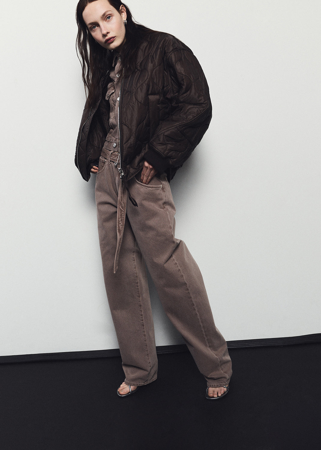 Person wearing a dark jacket and brown pants against a white wall.