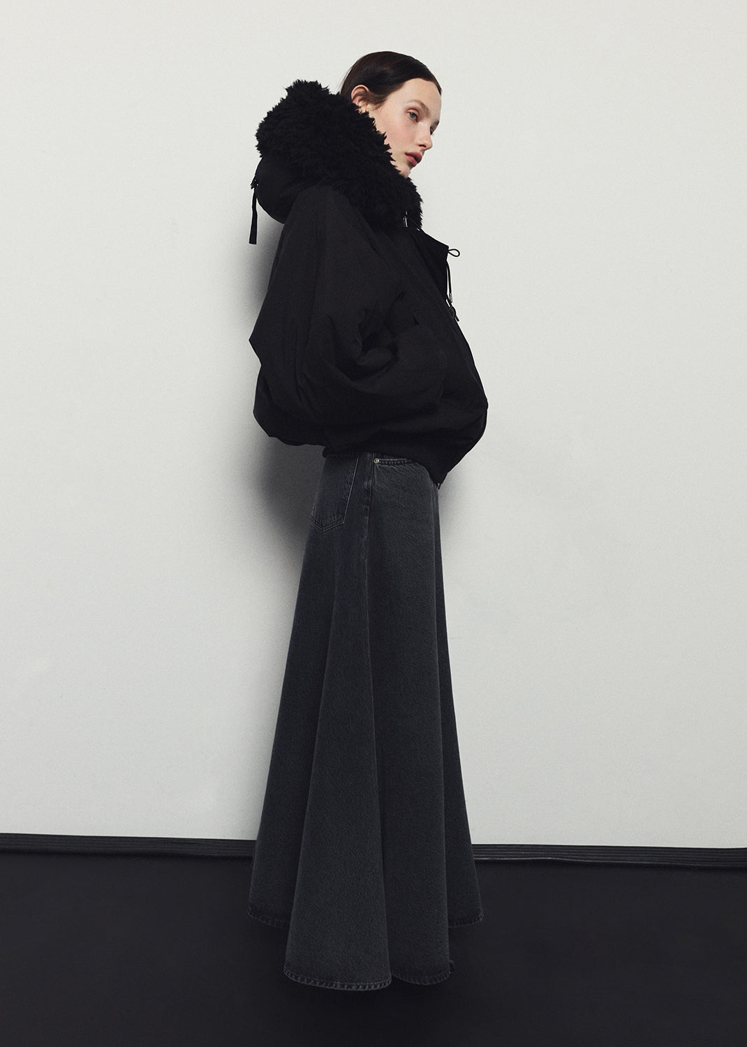 Person wearing a black hoodie and dark jeans against a white wall
