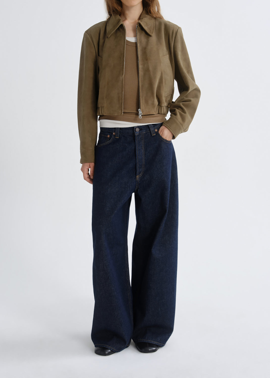 A model wearing a cropped olive-brown suede jacket, a beige top, white undershirt, and dark wide-leg denim jeans, standing against a grey background.