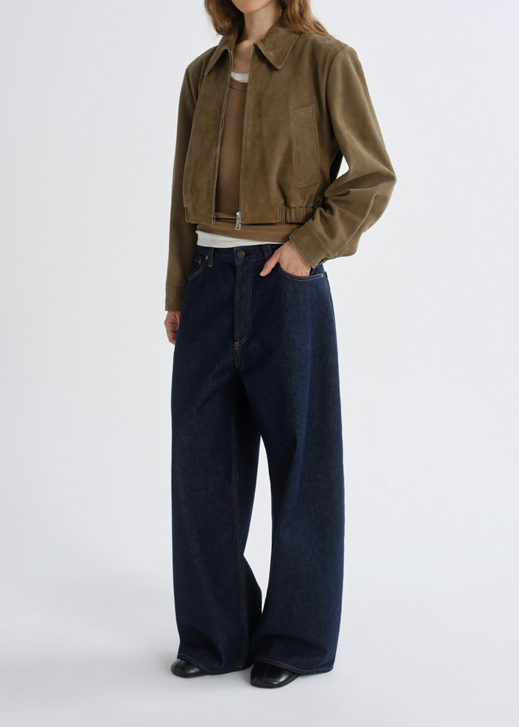 A model wearing a cropped olive-brown suede jacket, a white ribbed top, dark wide-leg denim jeans, and black shoes, standing against a grey background.