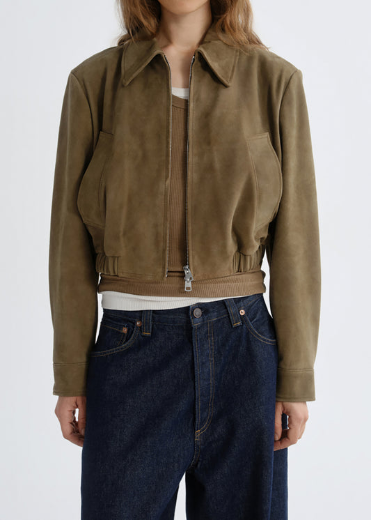 A cropped suede jacket in a muted olive-brown color, worn by a model with dark blue jeans and a white top visible underneath. The jacket features a front zipper, collar, and side pockets, photographed against a grey background.