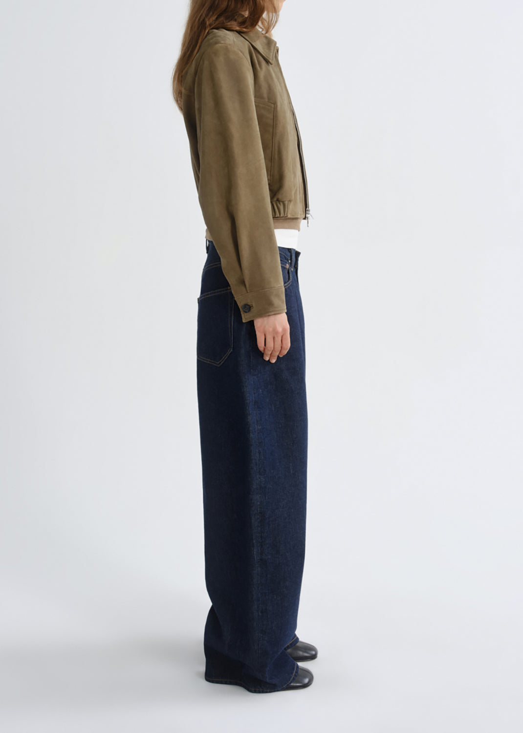 A model shown from the side wearing a cropped olive-brown suede jacket, dark blue wide-leg jeans, and black shoes against a grey background.