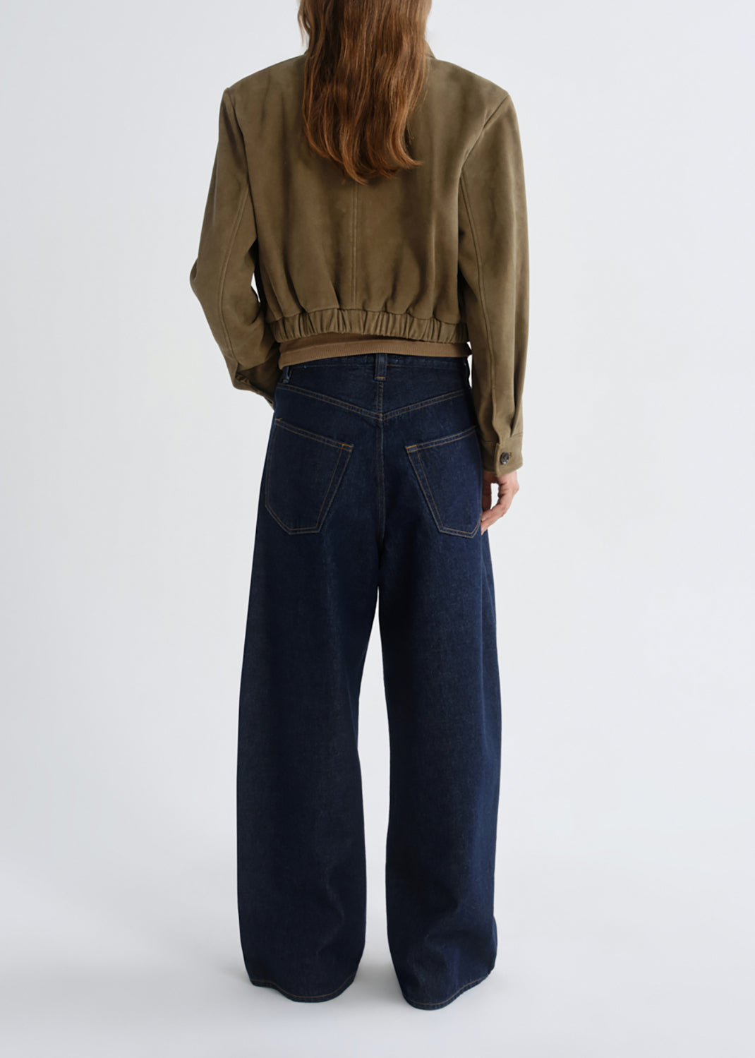 A model shown from the back wearing a cropped olive green jacket and dark wide-leg denim jeans against a grey background.