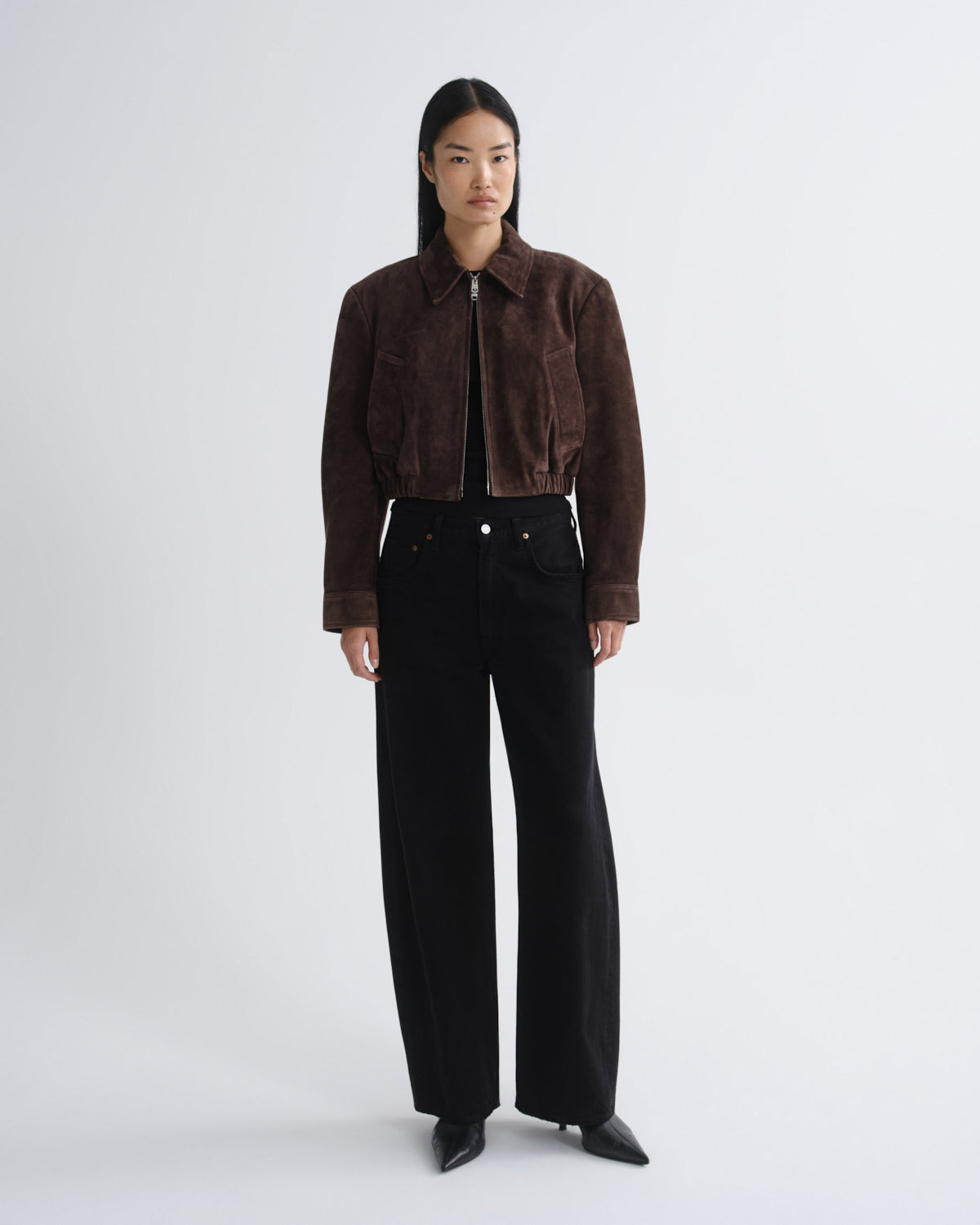 A model stands against a light gray studio background wearing a dark brown cropped suede jacket with a front zipper, black wide-leg pants, and black pointed shoes.