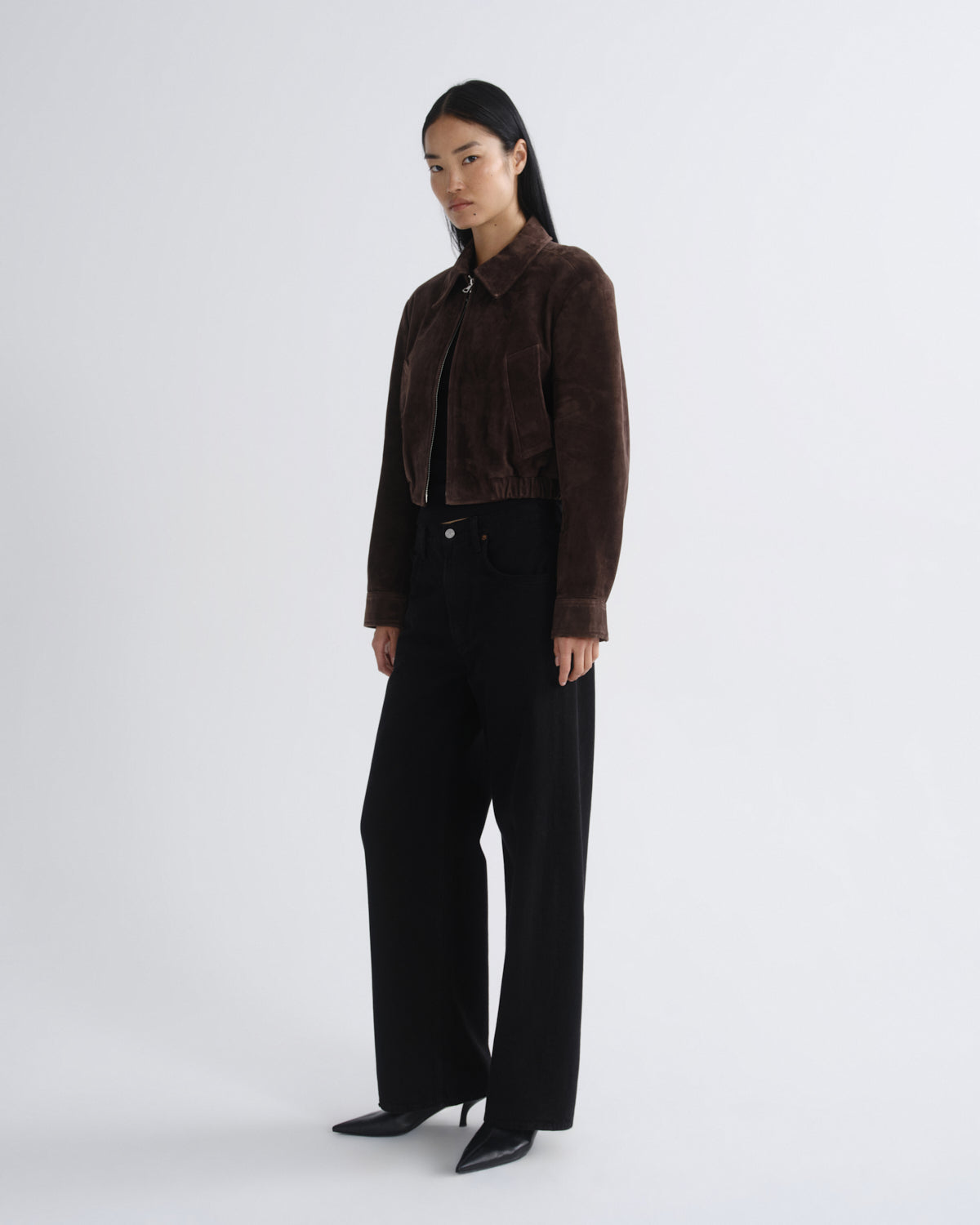 A model stands in a studio wearing a dark brown cropped suede jacket, black wide-leg trousers, and black pointed-toe heels against a light gray background.