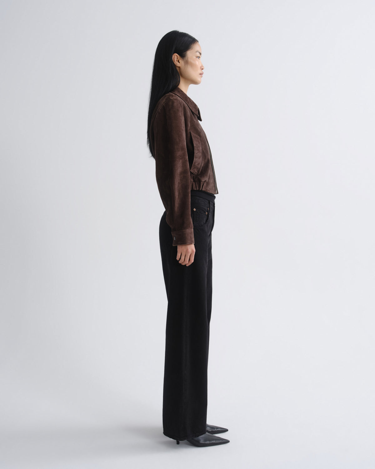 A woman stands in profile wearing a dark brown suede jacket and black wide-leg trousers against a plain light gray studio background.