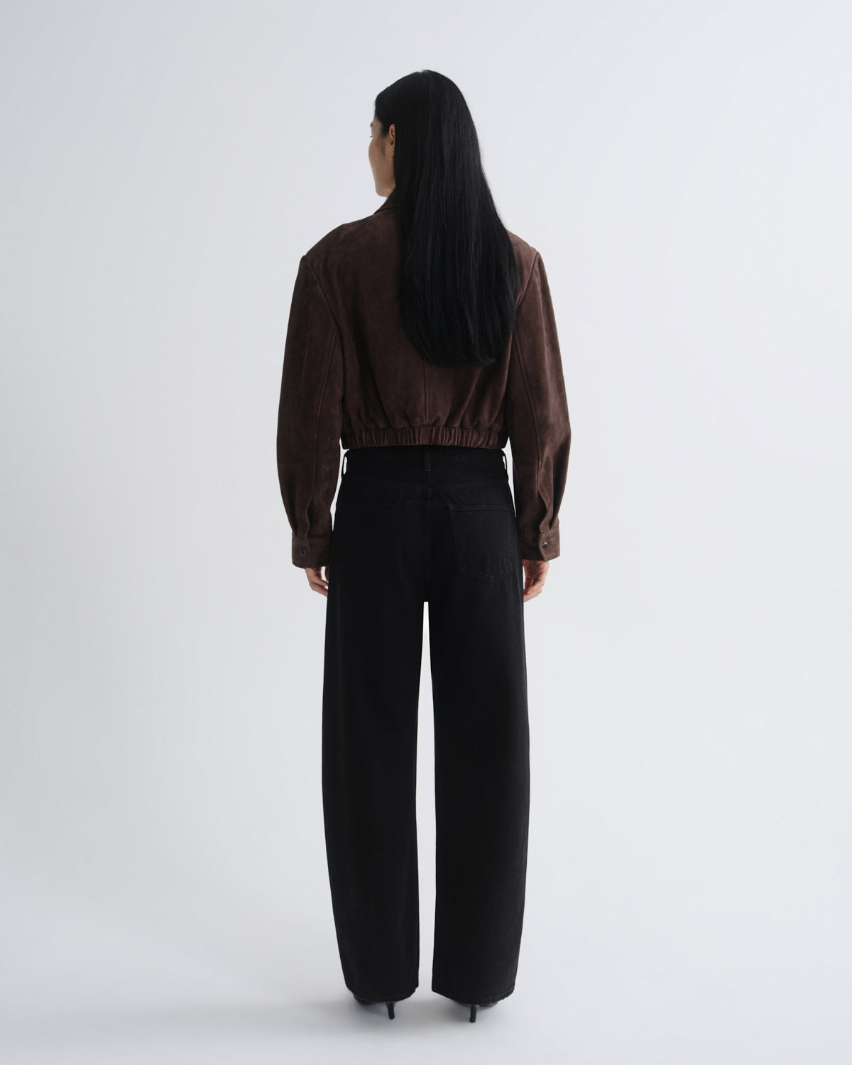 A model shown from the back wearing a dark brown cropped velvet jacket and black wide-leg trousers, standing against a light gray studio background.