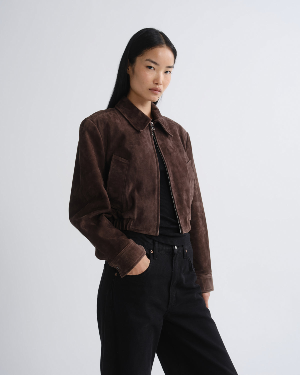 A model wearing a dark brown suede cropped jacket over a black top and black pants, standing against a light gray studio background.