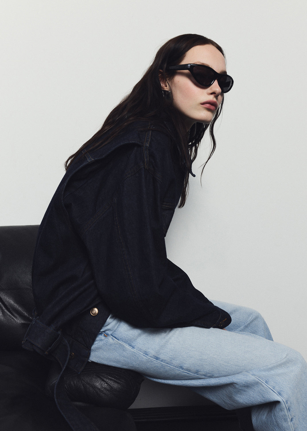 Person wearing a black coat and sunglasses sitting on a black chair against a white background