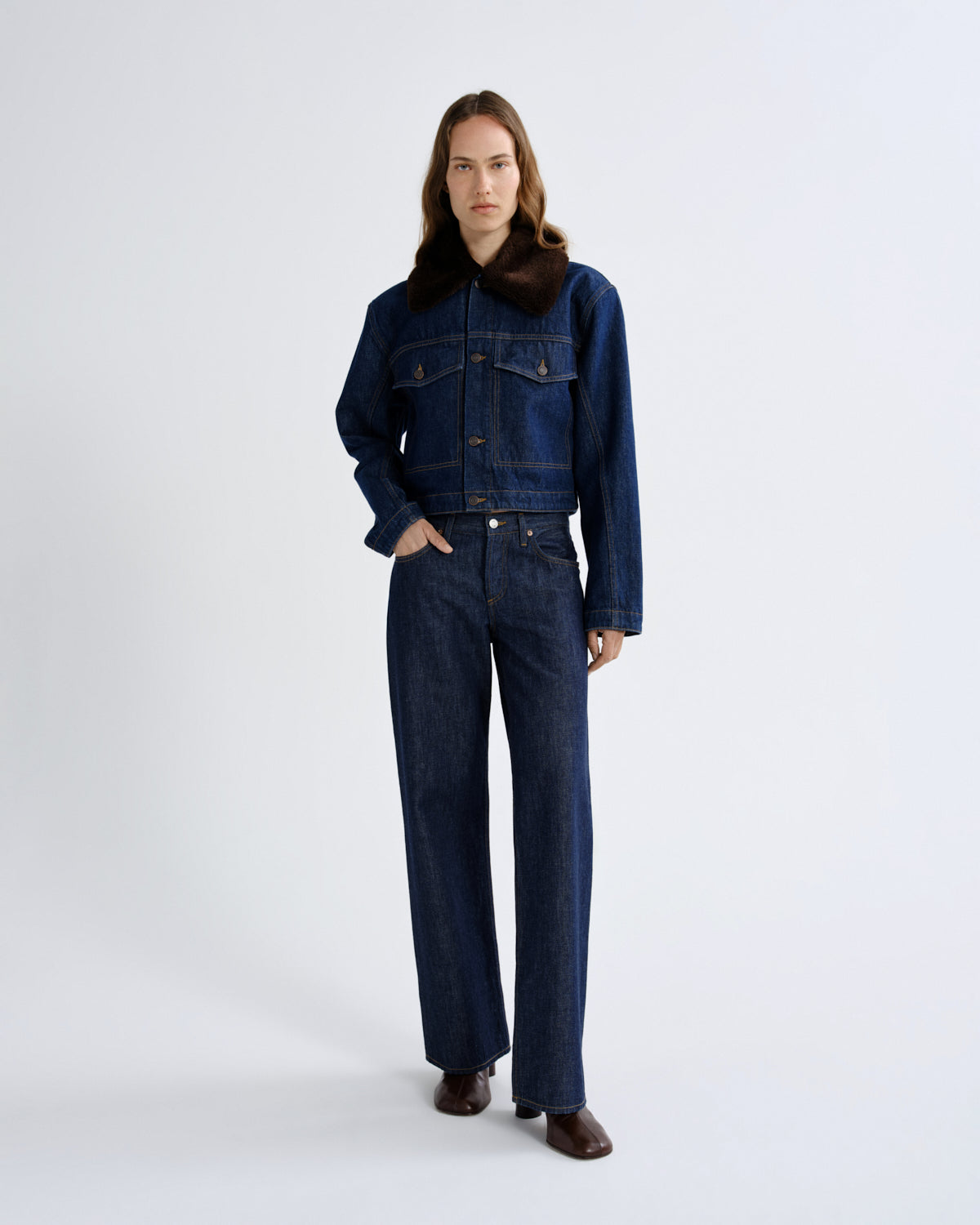 A model stands against a light gray studio background wearing a dark blue denim jacket with a brown collar and matching dark blue wide-leg denim pants.