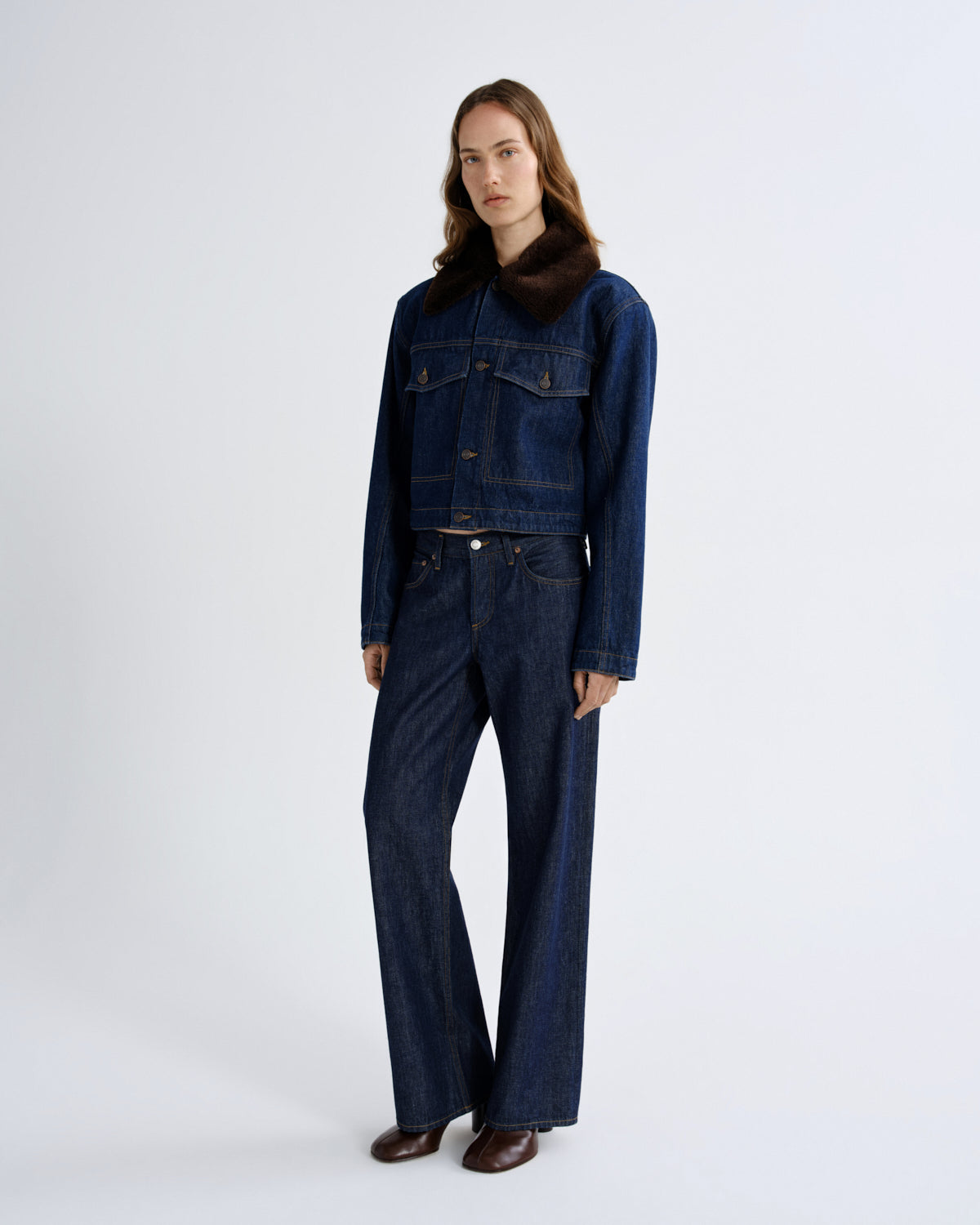 A model stands against a light gray studio background wearing a dark blue denim jacket with a dark brown fur collar, matching dark blue wide-leg denim pants, and brown leather shoes.