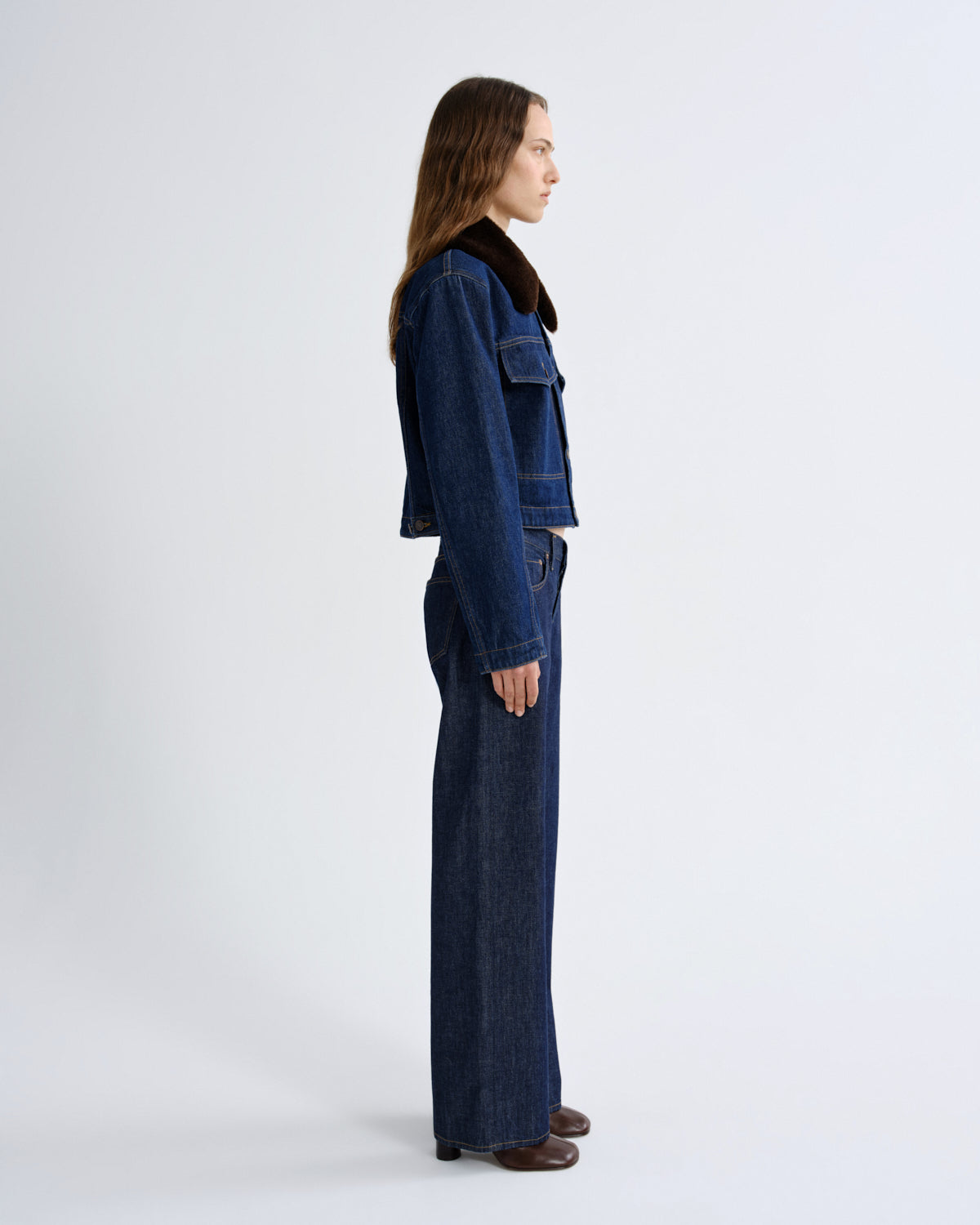A woman shown in profile wearing a dark blue denim jacket with a brown shearling-style collar and matching wide-leg denim pants, photographed against a clean light gray studio background.