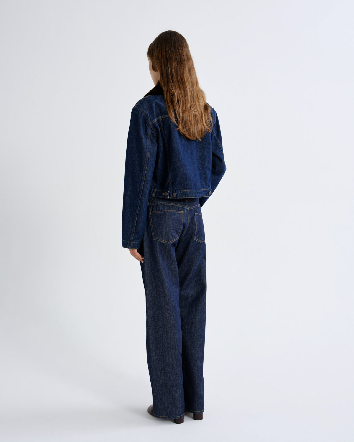 A model viewed from the back wearing a dark blue cropped denim jacket and matching wide-leg denim pants against a plain white studio background.