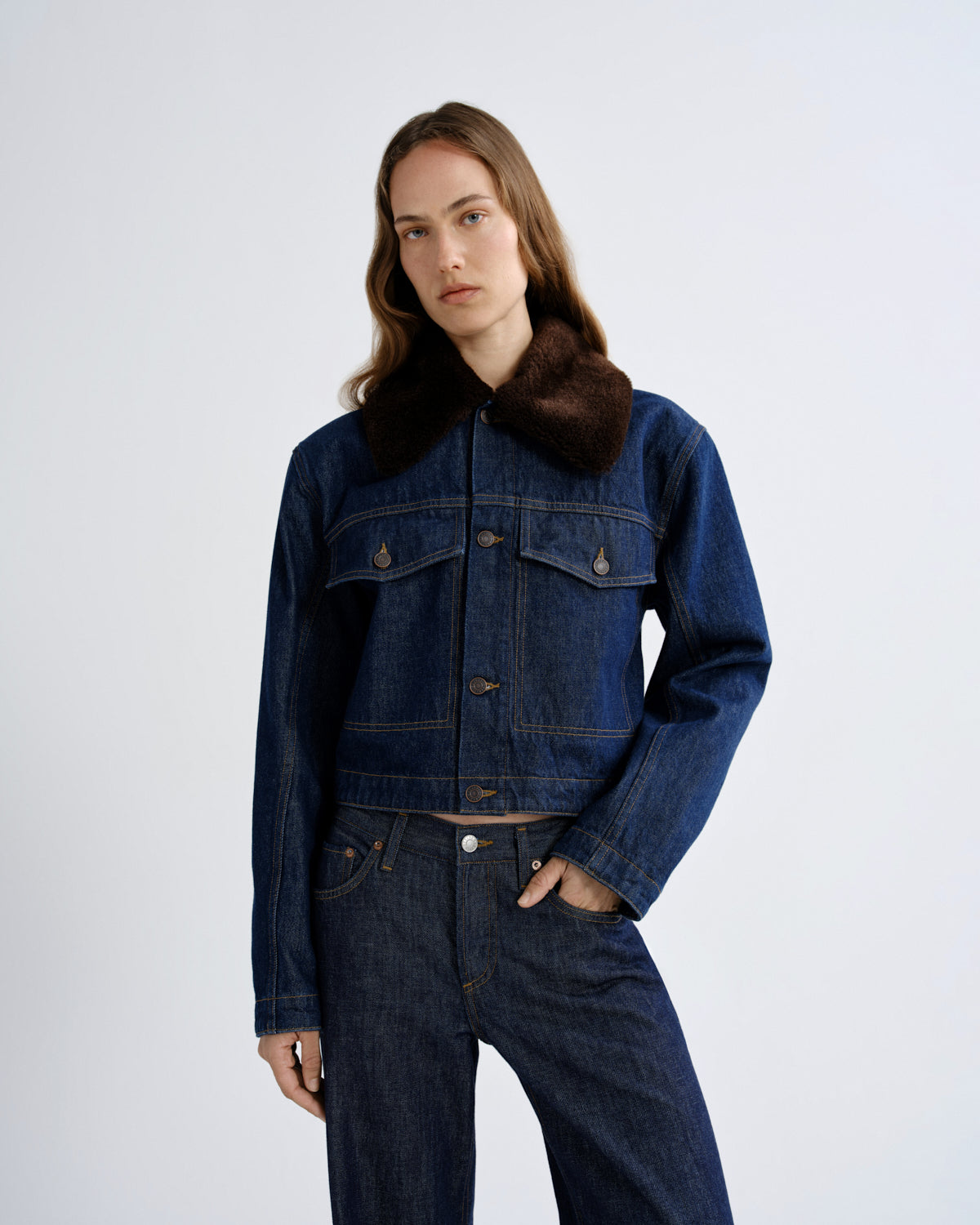 A model wearing a cropped dark blue denim jacket with a thick brown faux-fur collar, paired with dark blue jeans, against a light gray studio background.