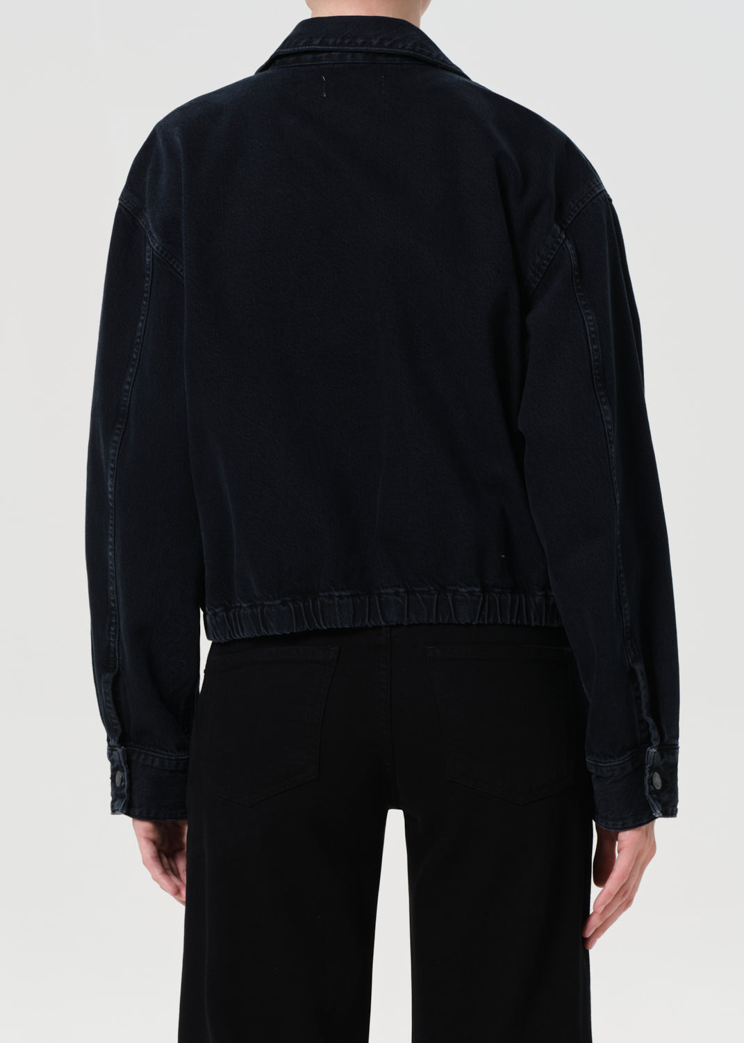 Person wearing a blue denim jacket over a white shirt and black pants on a light gray background