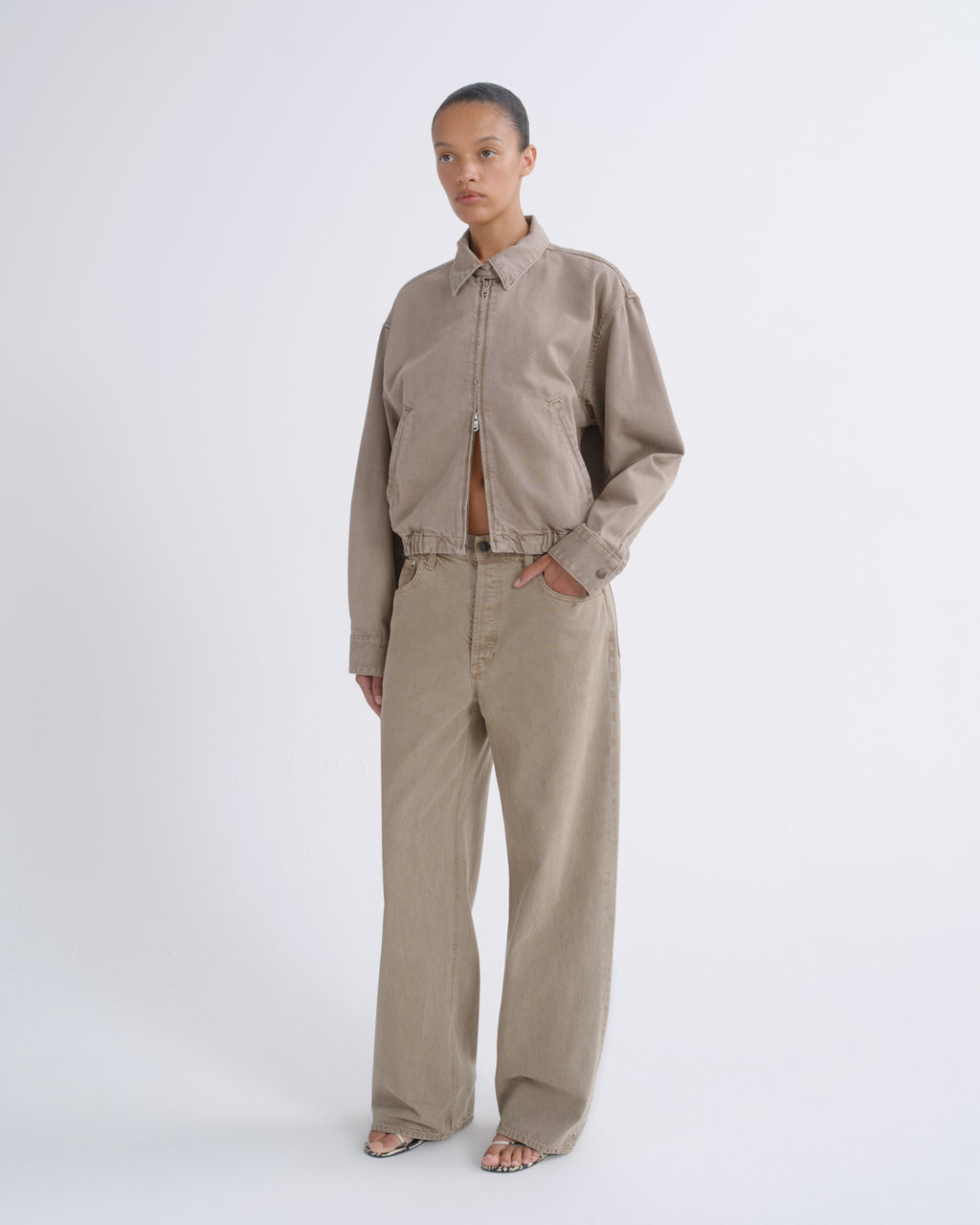 A model stands against a light gray studio background wearing a cropped zip-front jacket and matching wide-leg trousers in a warm taupe-beige color.