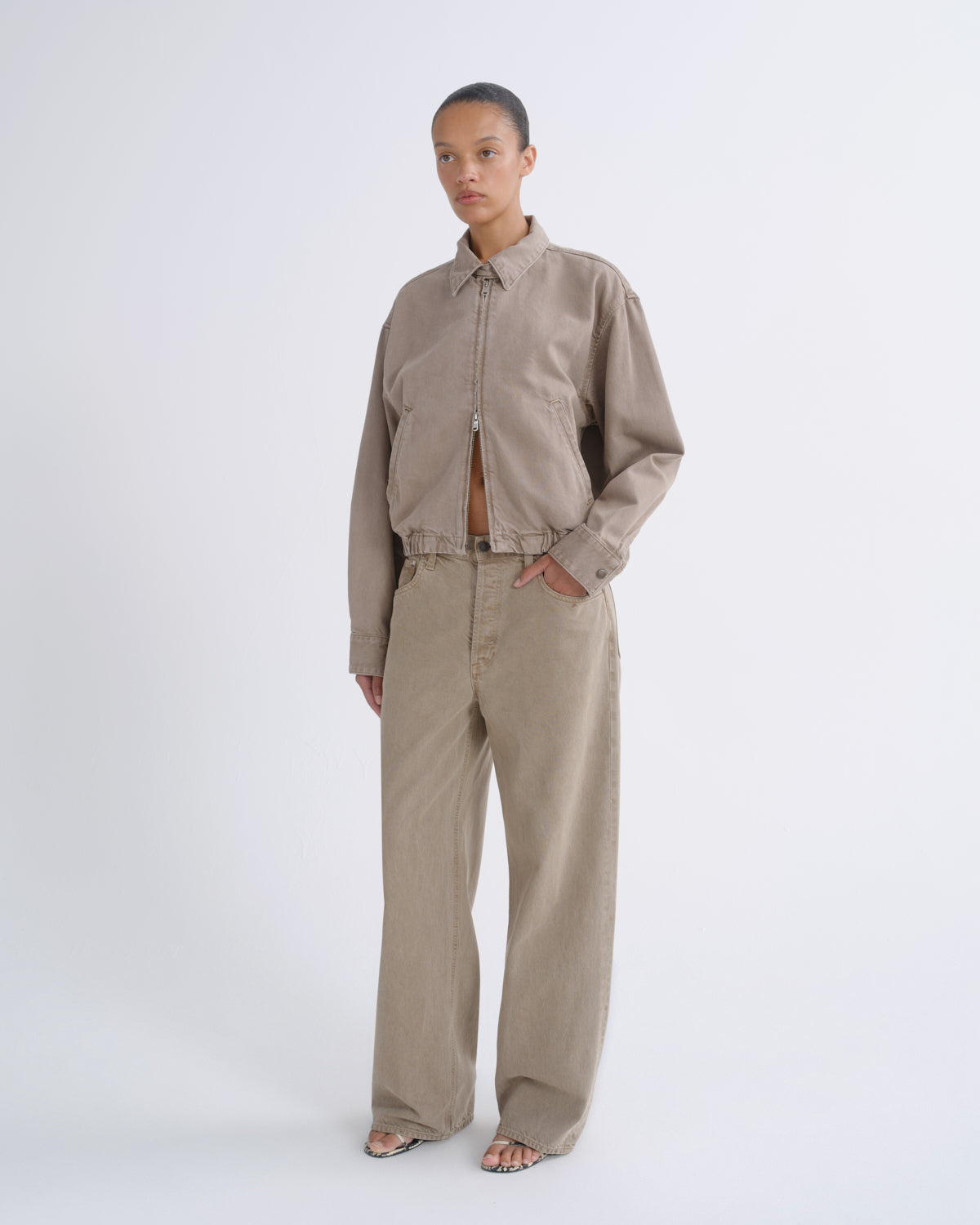 A model stands against a light gray studio background wearing a cropped zip-front jacket and matching wide-leg trousers in a warm taupe-beige color.