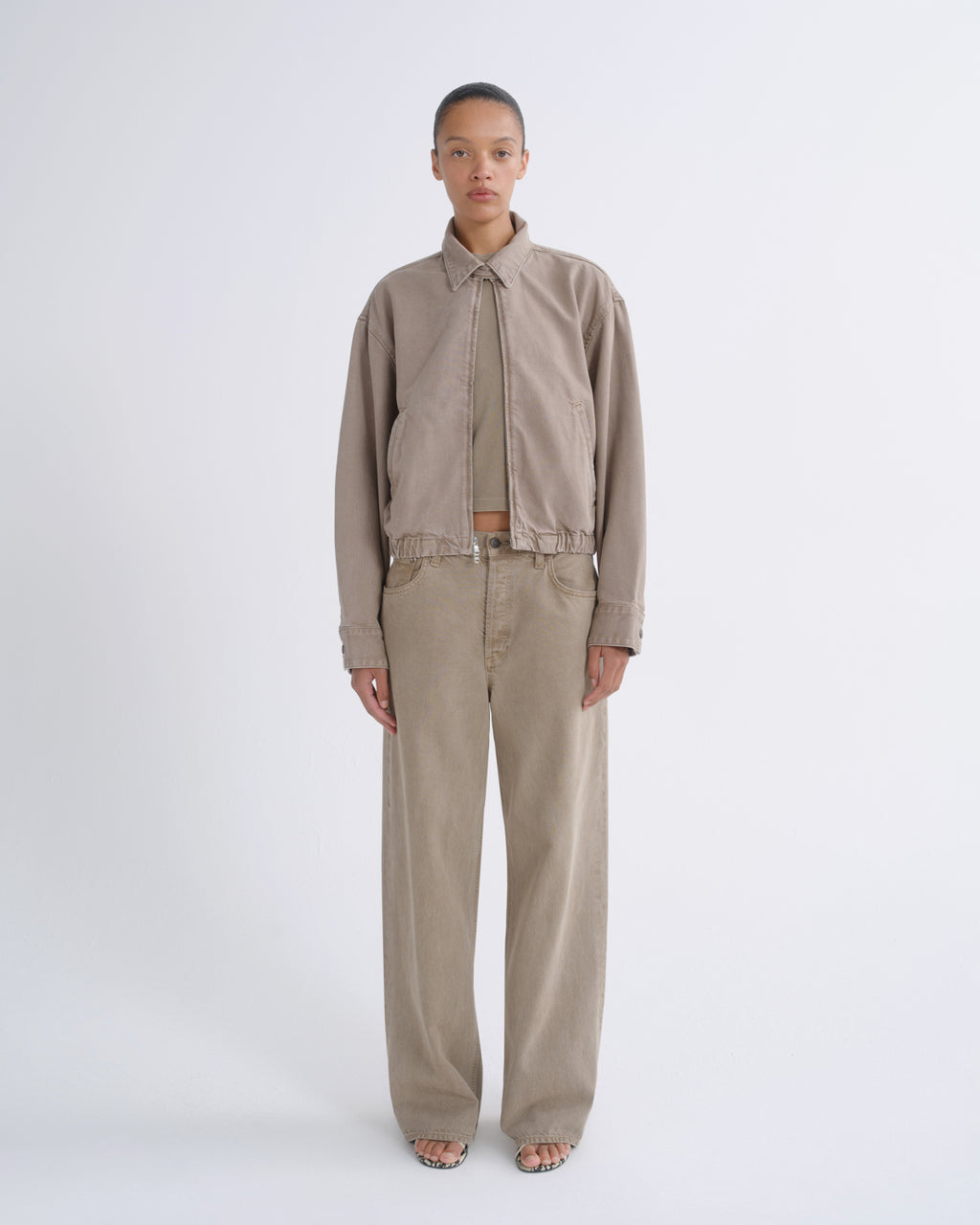 A model stands against a light gray studio background wearing a beige cropped button-up shirt with a tie-neck detail, matching beige wide-leg pants, and a thin belt.