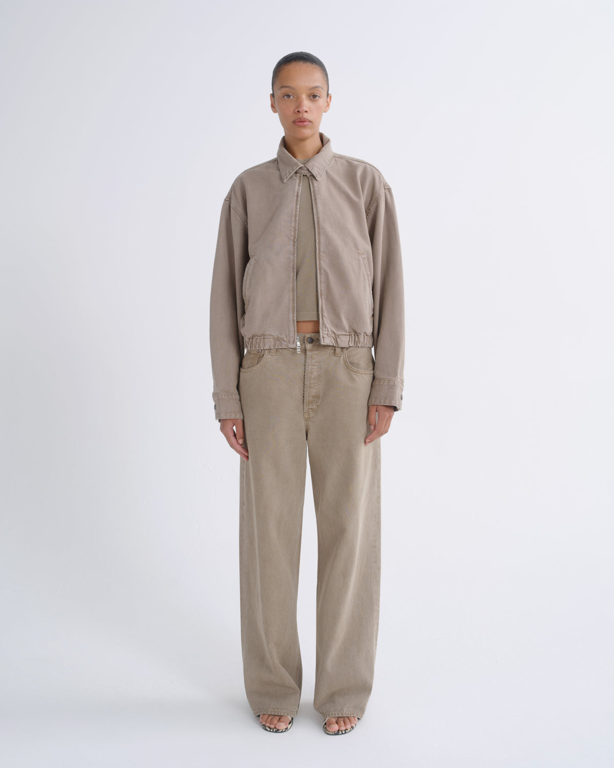 A model stands against a light gray studio background wearing a beige cropped button-up shirt with a tie-neck detail, matching beige wide-leg pants, and a thin belt.