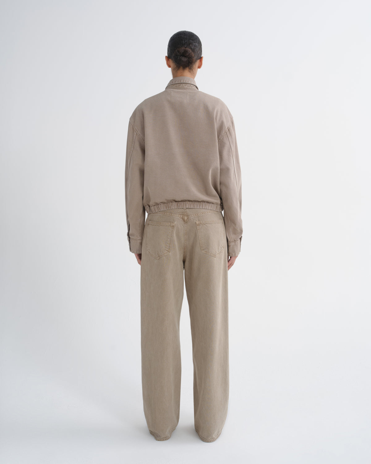 A model viewed from the back wearing a beige long-sleeve cropped jacket and matching beige wide-leg trousers against a plain light gray studio background.