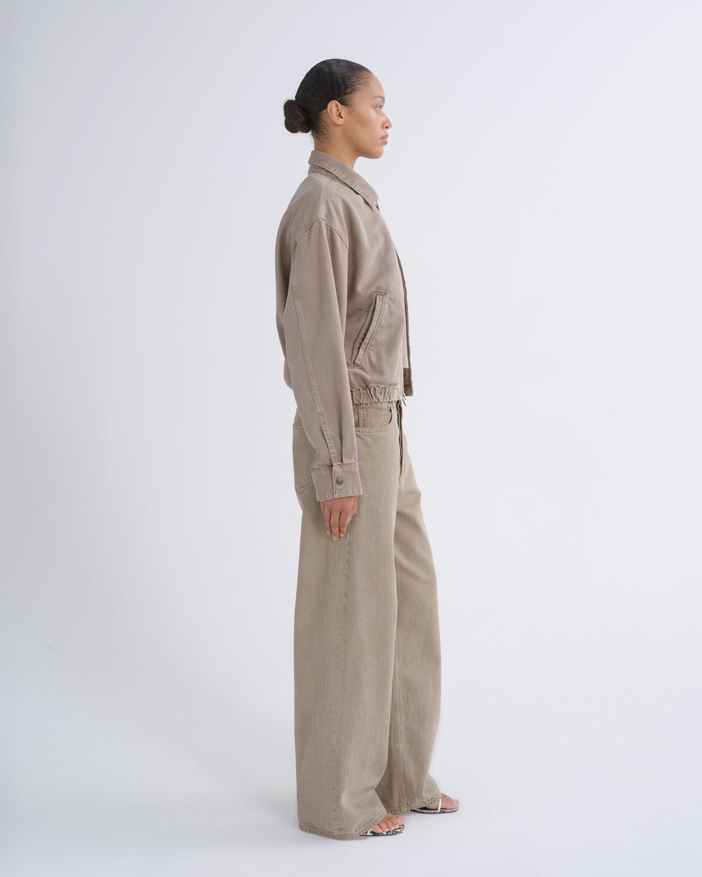 A model in side profile wearing a matching beige linen-look jacket and wide-leg trousers set against a plain light gray studio background.