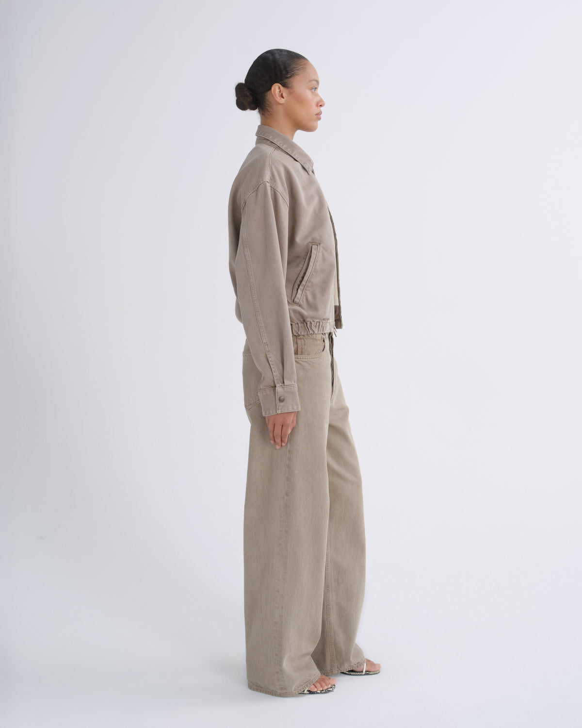 A model in side profile wearing a matching beige linen-look jacket and wide-leg trousers set against a plain light gray studio background.