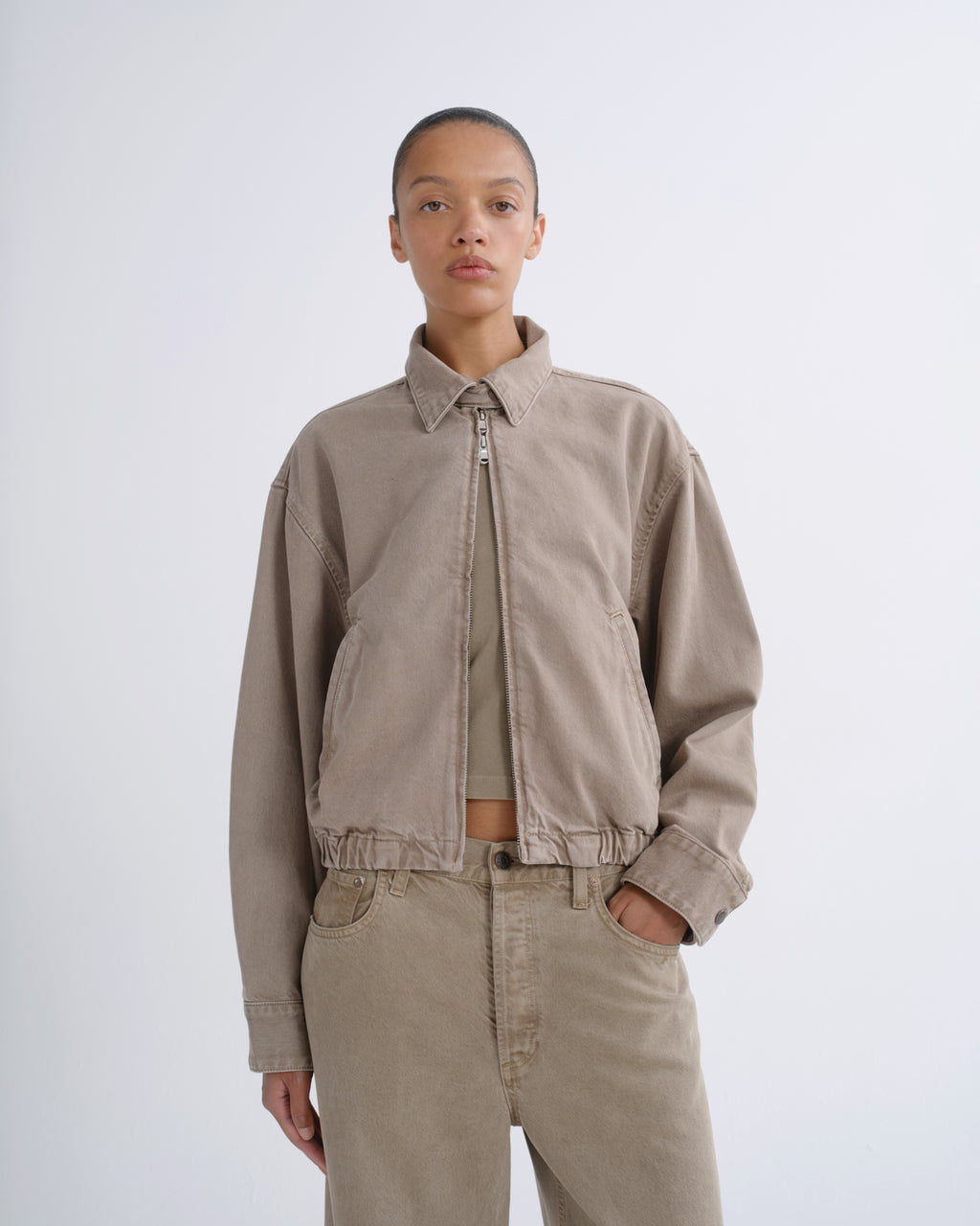 A model wearing a light beige cropped utility jacket with a zip-up front, paired with matching beige trousers and a beige top, shot against a plain light gray studio background.