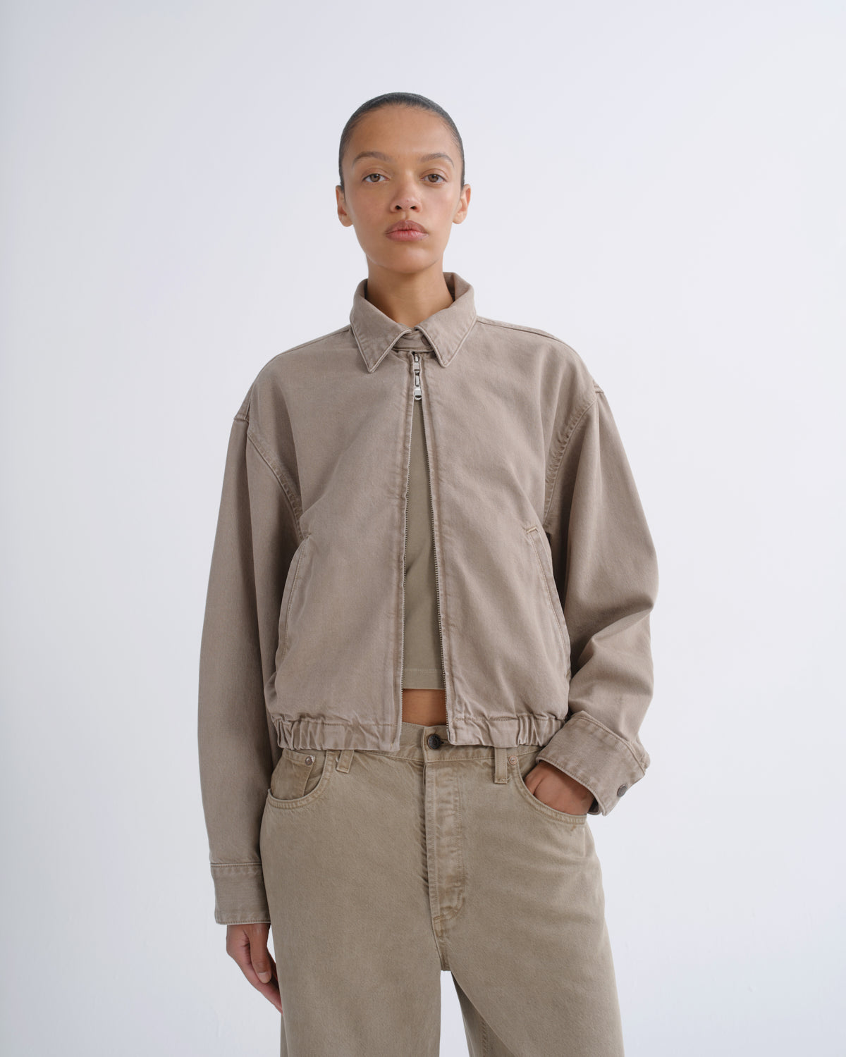 A model wearing a light beige cropped utility jacket with a zip-up front, paired with matching beige trousers and a beige top, shot against a plain light gray studio background.