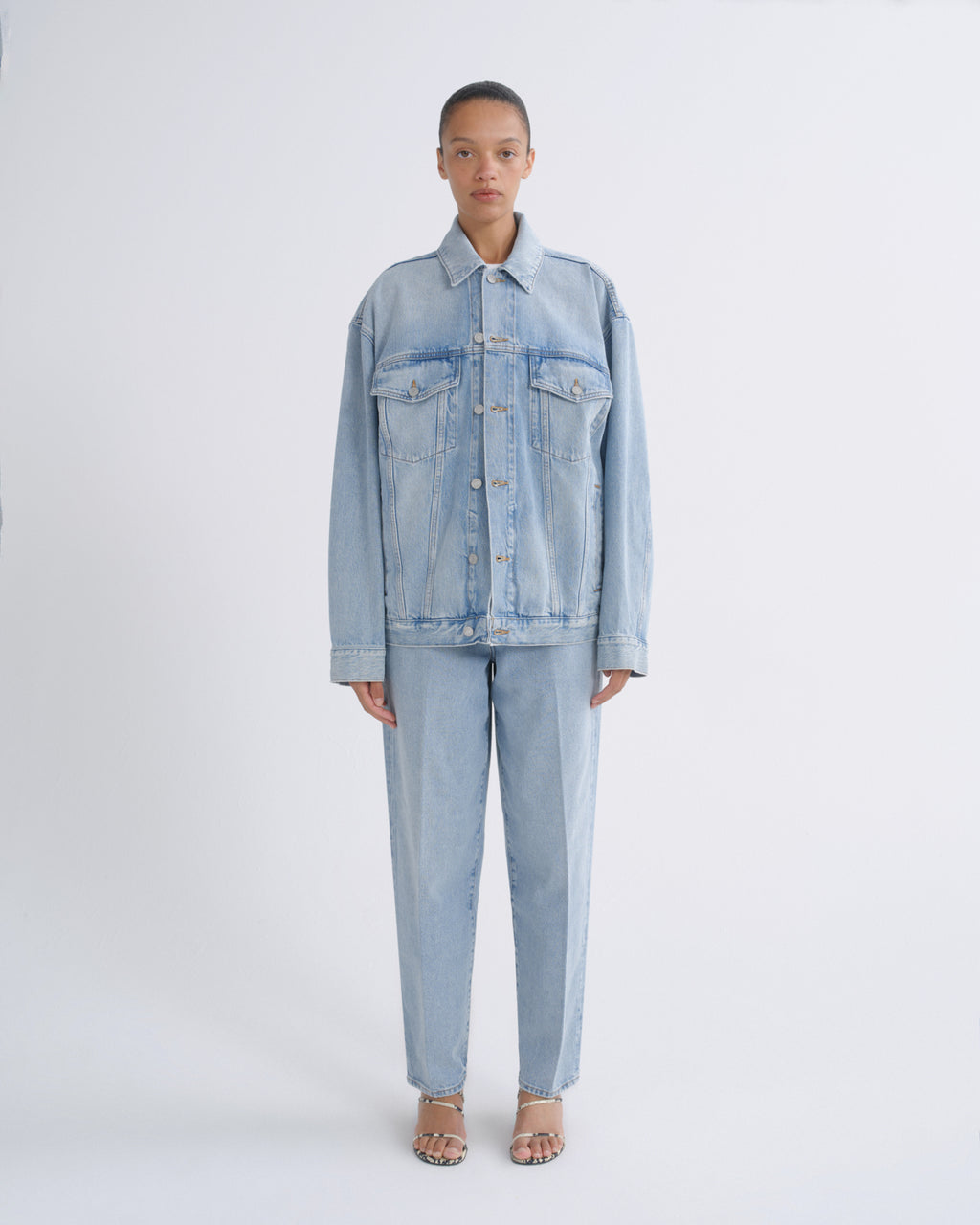 A model stands against a light gray studio background wearing a light blue denim jacket and matching light blue denim pants.