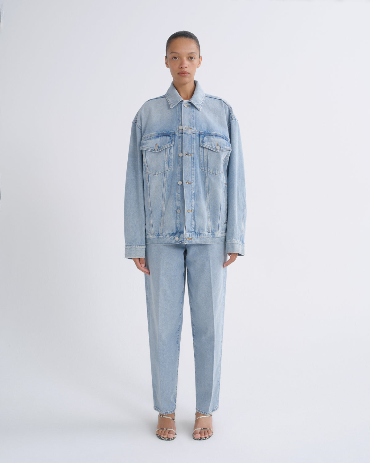 A model stands against a light gray studio background wearing a light blue denim jacket and matching light blue denim pants.