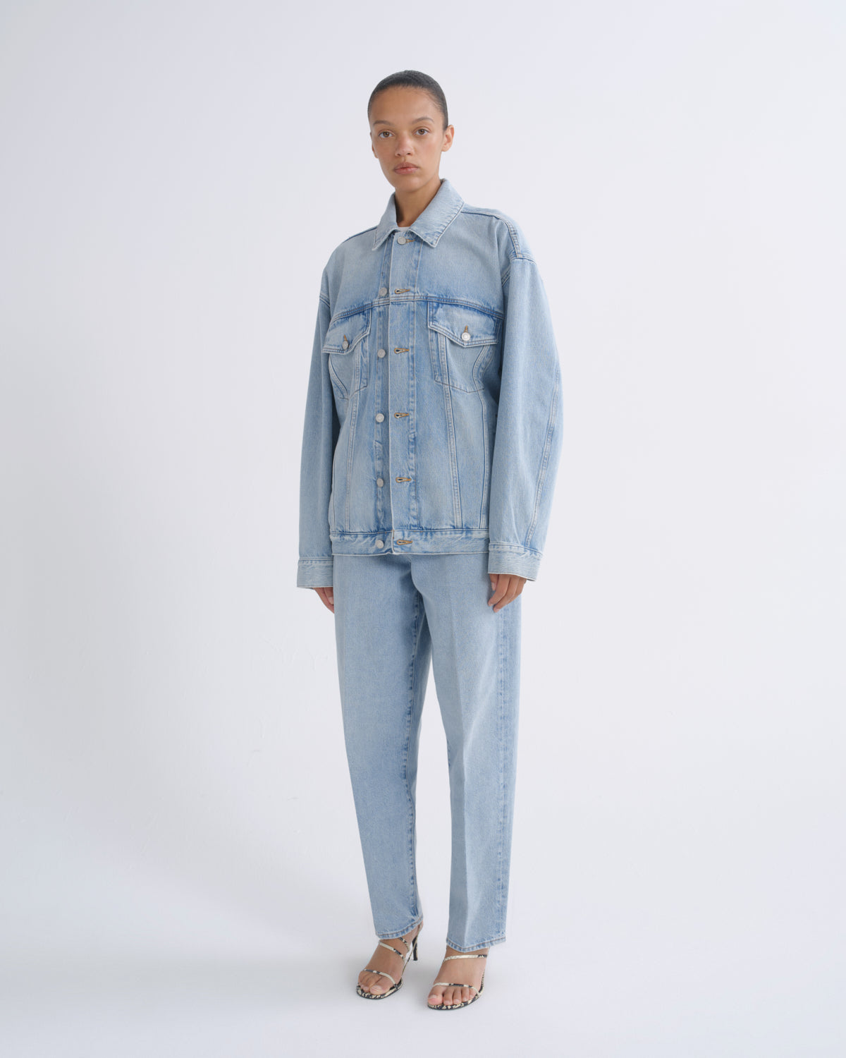 A model stands against a light gray studio background wearing a light-wash blue denim jacket and matching light-wash blue denim jeans, paired with metallic heeled sandals.