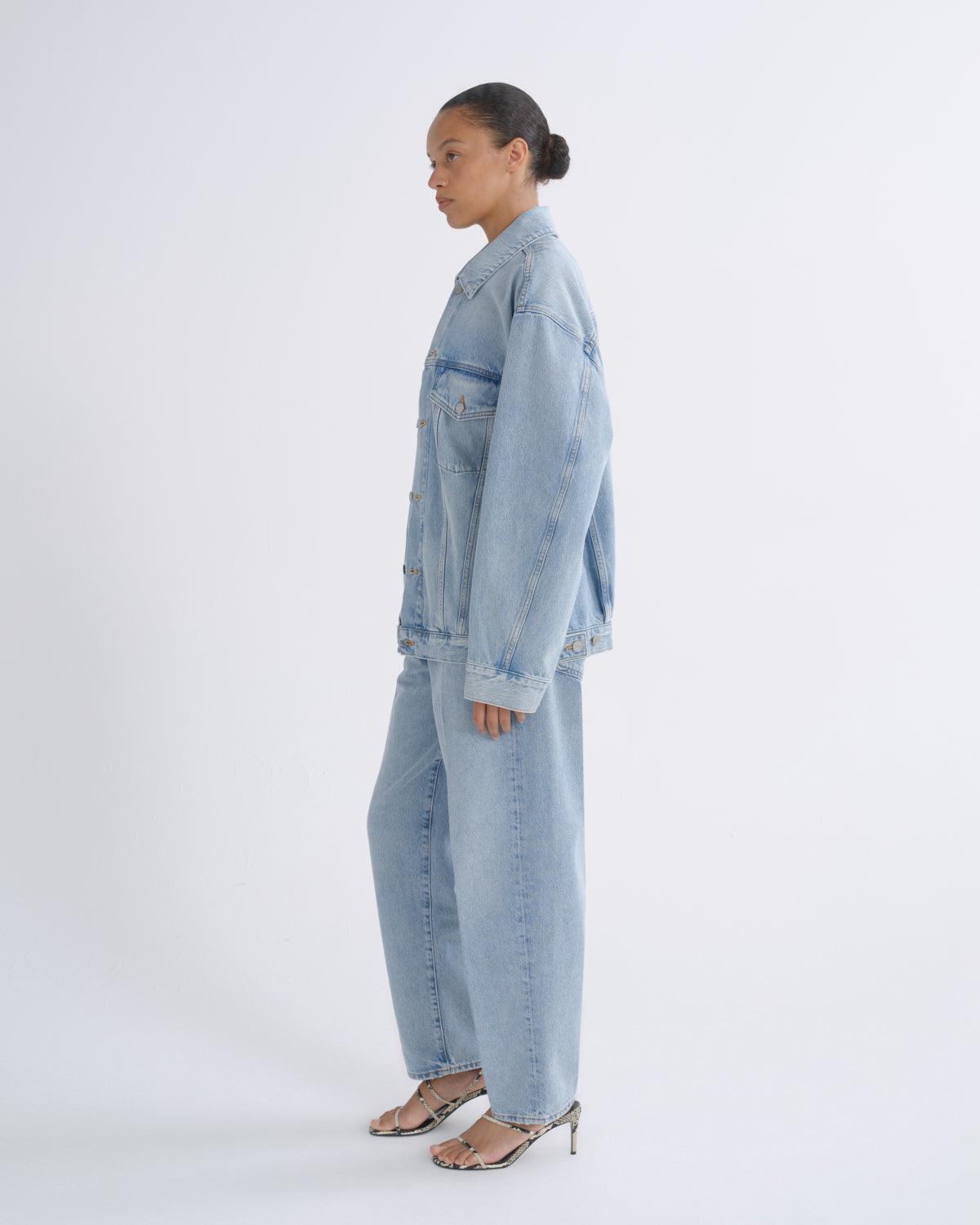 A model shown in profile wearing a matching light-wash denim set consisting of a cropped denim jacket and wide-leg denim pants, paired with metallic heeled sandals, against a plain white studio background.