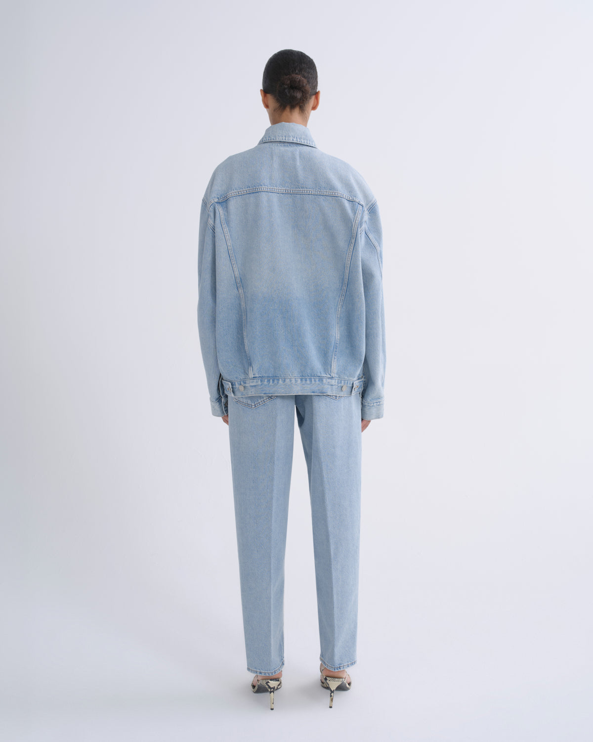 A model viewed from behind wearing a light blue denim jacket and matching light blue denim trousers, paired with metallic high heels, against a plain light gray studio background.