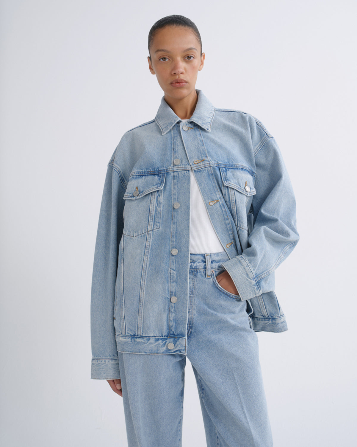 A model wearing a light-wash blue denim jacket and matching wide-leg jeans over a white t-shirt, photographed against a neutral light gray studio background.