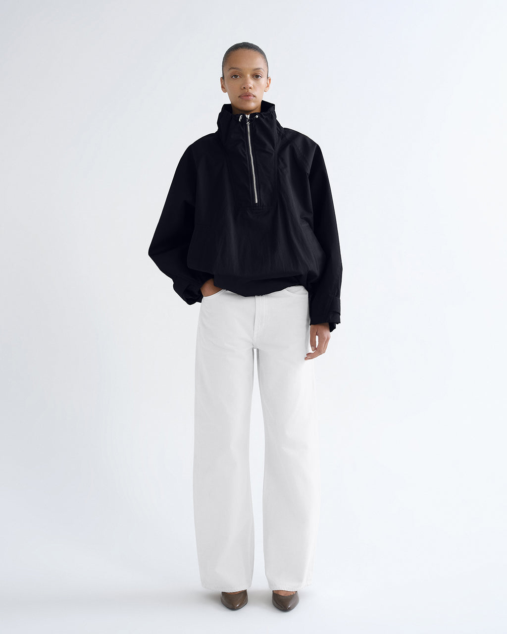 A model stands against a plain white studio background wearing a black cropped half-zip jacket with a high collar, white wide-leg trousers, and dark pointed shoes.