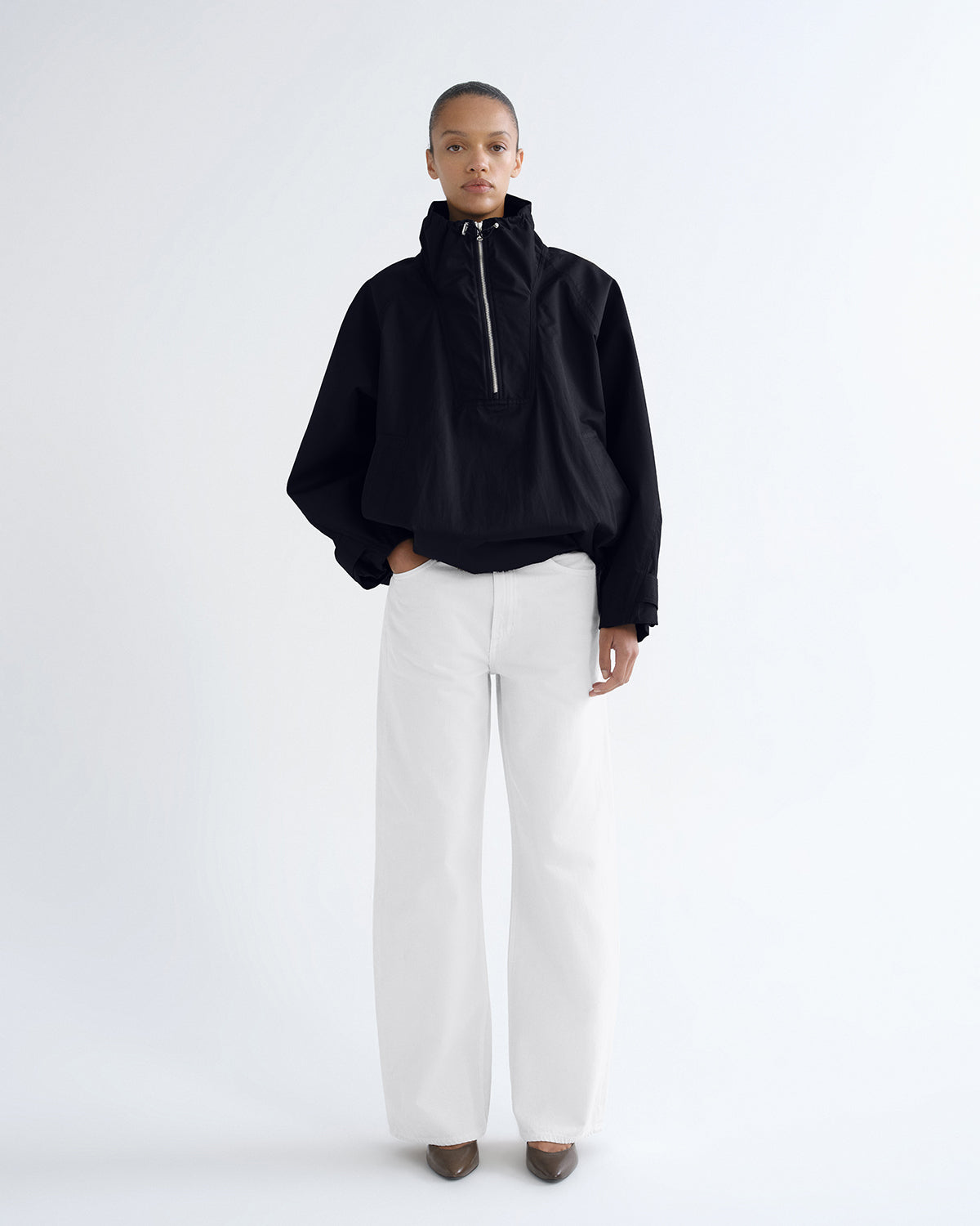 A model stands against a plain white studio background wearing a black cropped half-zip jacket with a high collar, white wide-leg trousers, and dark pointed shoes.