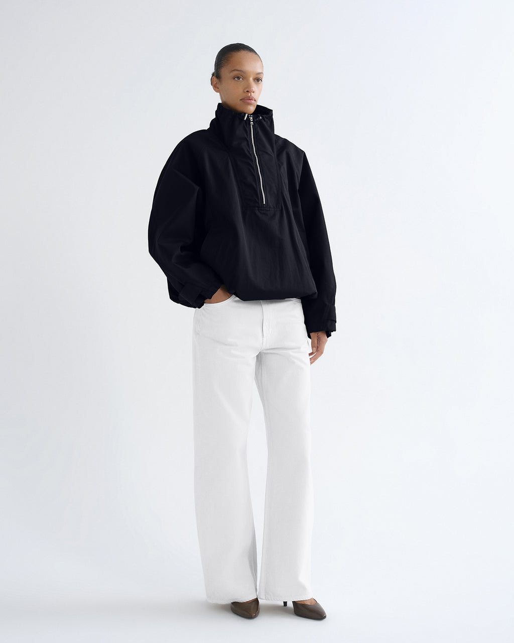 A model stands against a plain white studio background wearing a black half-zip pullover jacket with voluminous sleeves, paired with wide-leg white trousers and dark brown pointed shoes.
