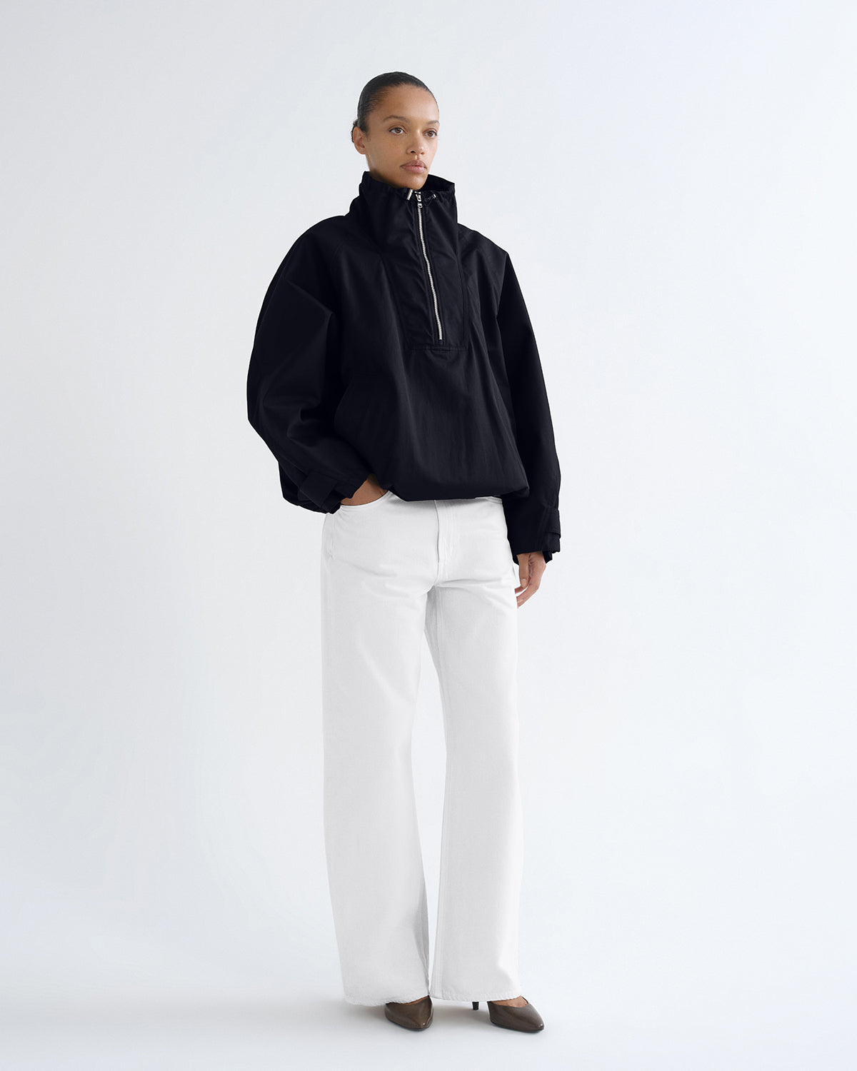 A model stands against a plain white studio background wearing a black half-zip pullover jacket with voluminous sleeves, paired with wide-leg white trousers and dark brown pointed shoes.
