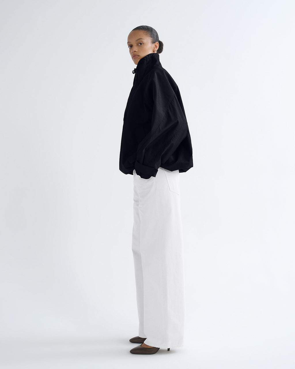 A model stands in profile wearing a black high-neck, oversized long-sleeve top and white wide-leg trousers against a plain white studio background.