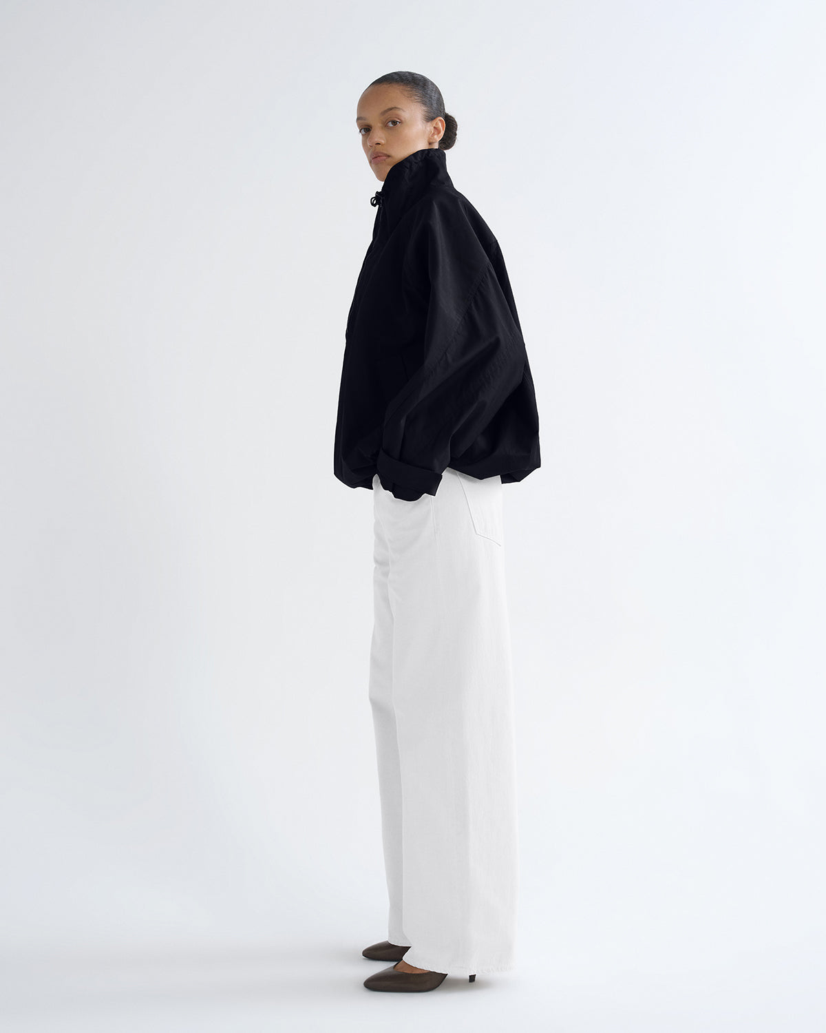 A model stands in profile wearing a black high-neck, oversized long-sleeve top and white wide-leg trousers against a plain white studio background.