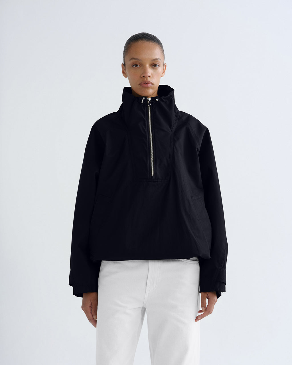 A model wearing a black cropped half-zip jacket with a high collar and white pants, standing against a light gray studio background.