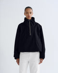 A model wearing a black cropped half-zip jacket with a high collar and white pants, standing against a light gray studio background.