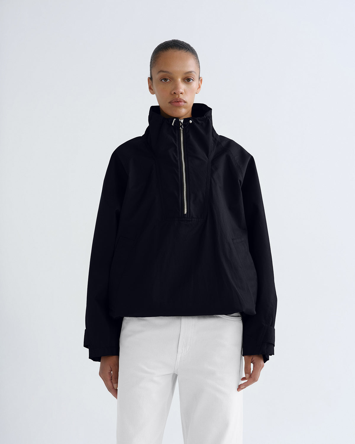 A model wearing a black cropped half-zip jacket with a high collar and white pants, standing against a light gray studio background.
