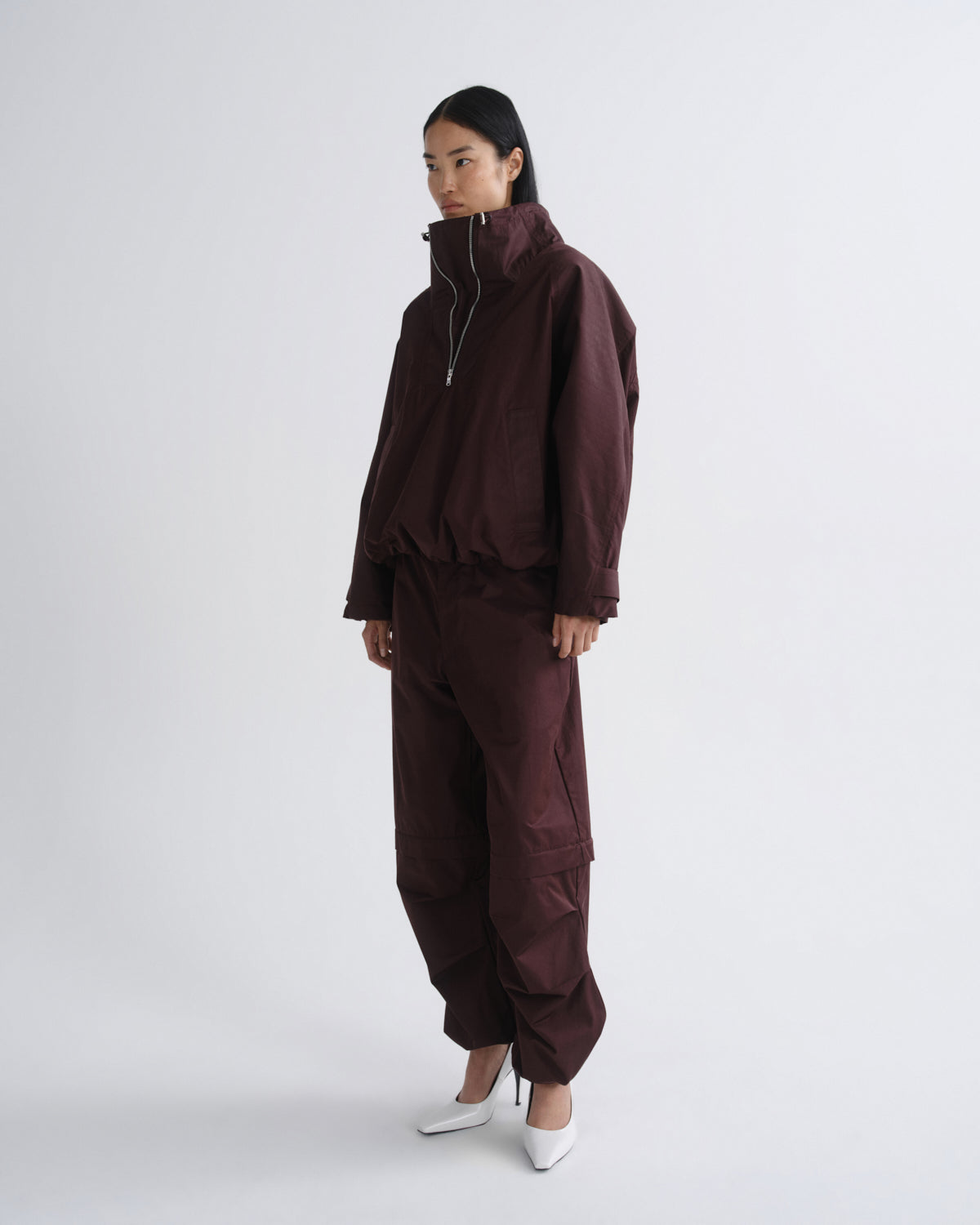 A model stands in profile wearing a matching dark burgundy zip-front jacket and cargo pants set, paired with white pointed heels, against a plain light gray studio background.